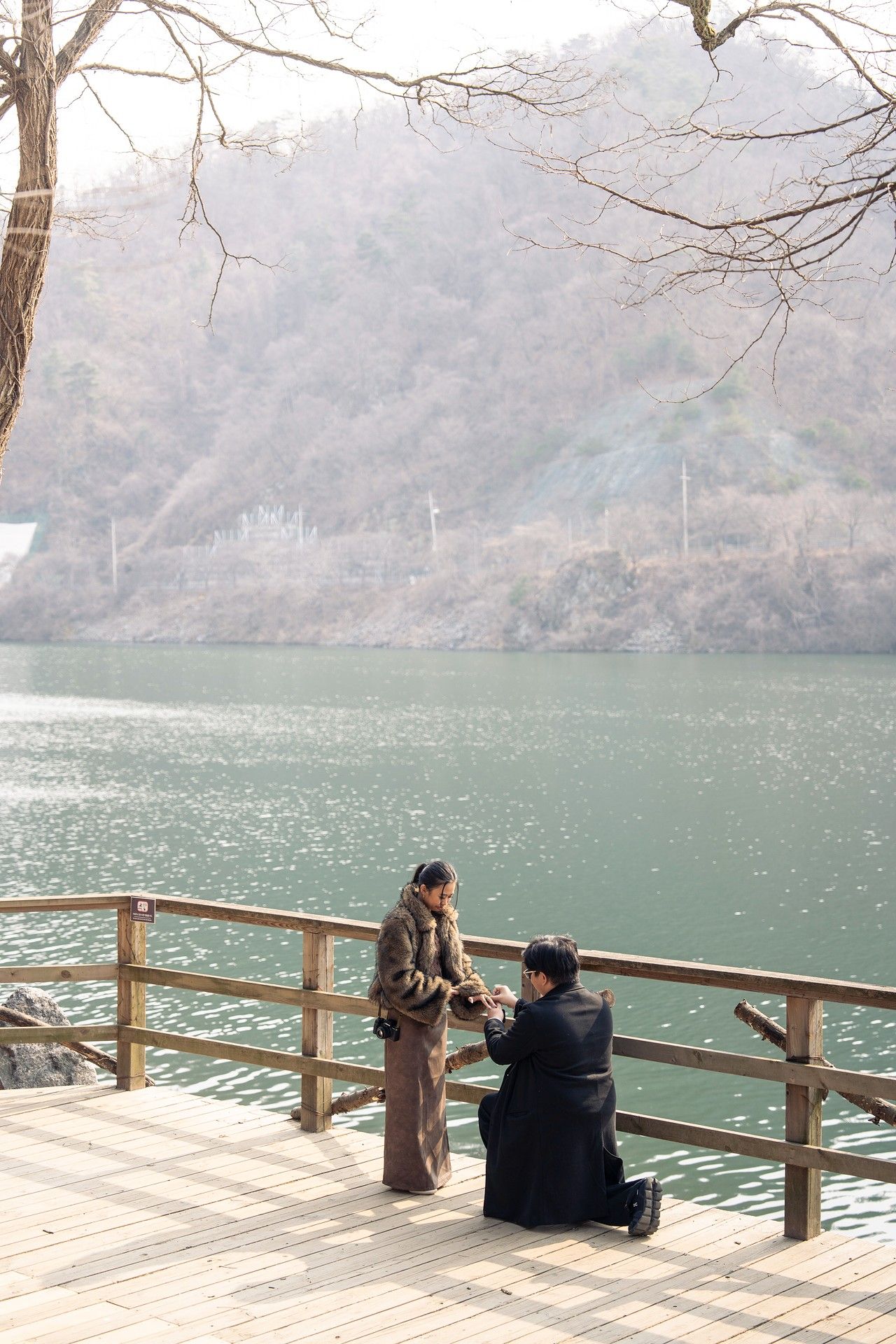 Man proposing to woman on wooden lakeside deck with scenic green water and misty mountain backdrop.