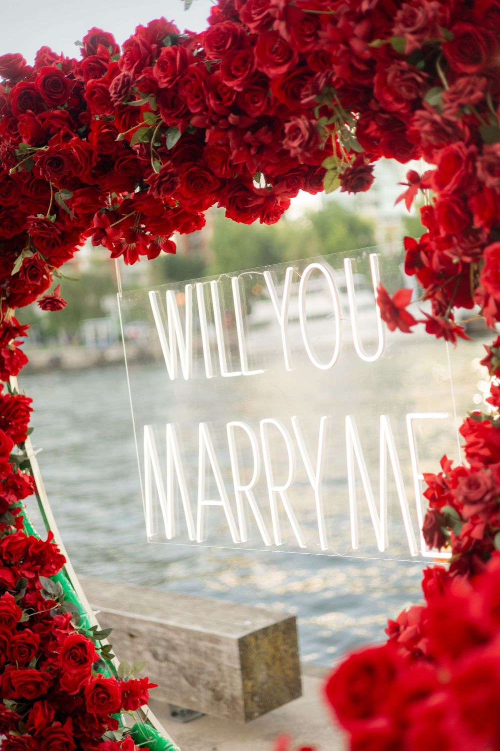 Heart-shaped red rose arch with Will You Marry Me neon sign beside a waterfront.