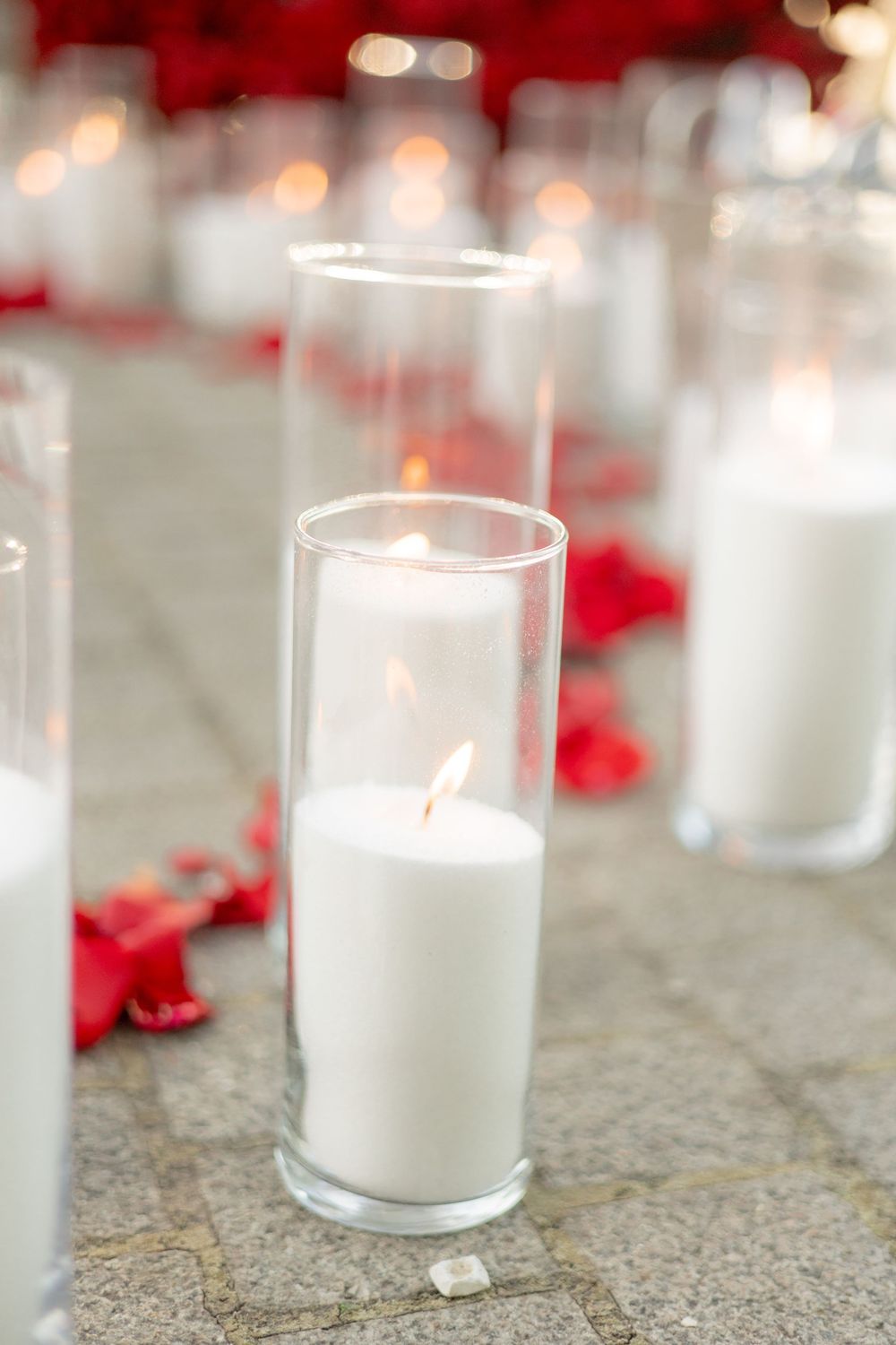 White pillar candles in glass cylinders on stone ground surrounded by red rose petals.