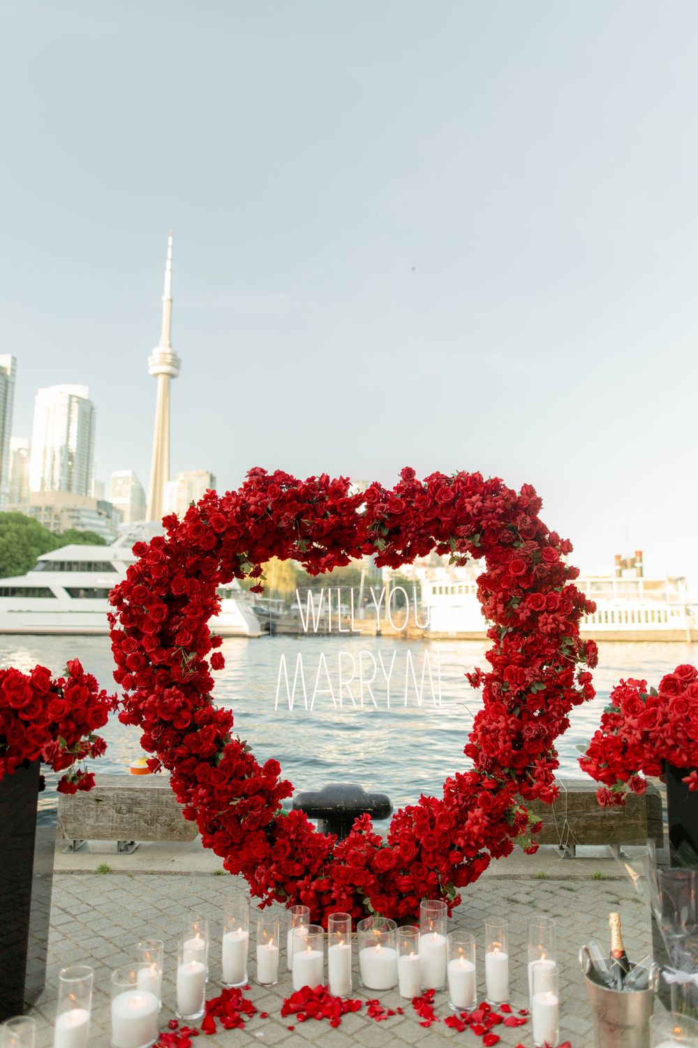 Romantic marriage proposal setup with red rose heart arch and Will You Marry Me sign by Toronto waterfront.