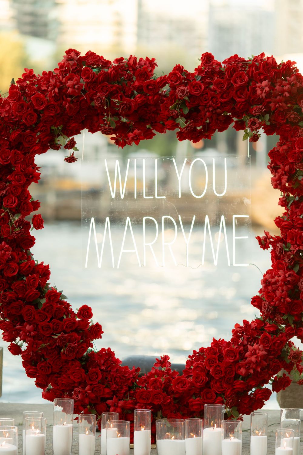 Close-up of heart-shaped red rose floral arch with Will You Marry Me neon sign and white candles.