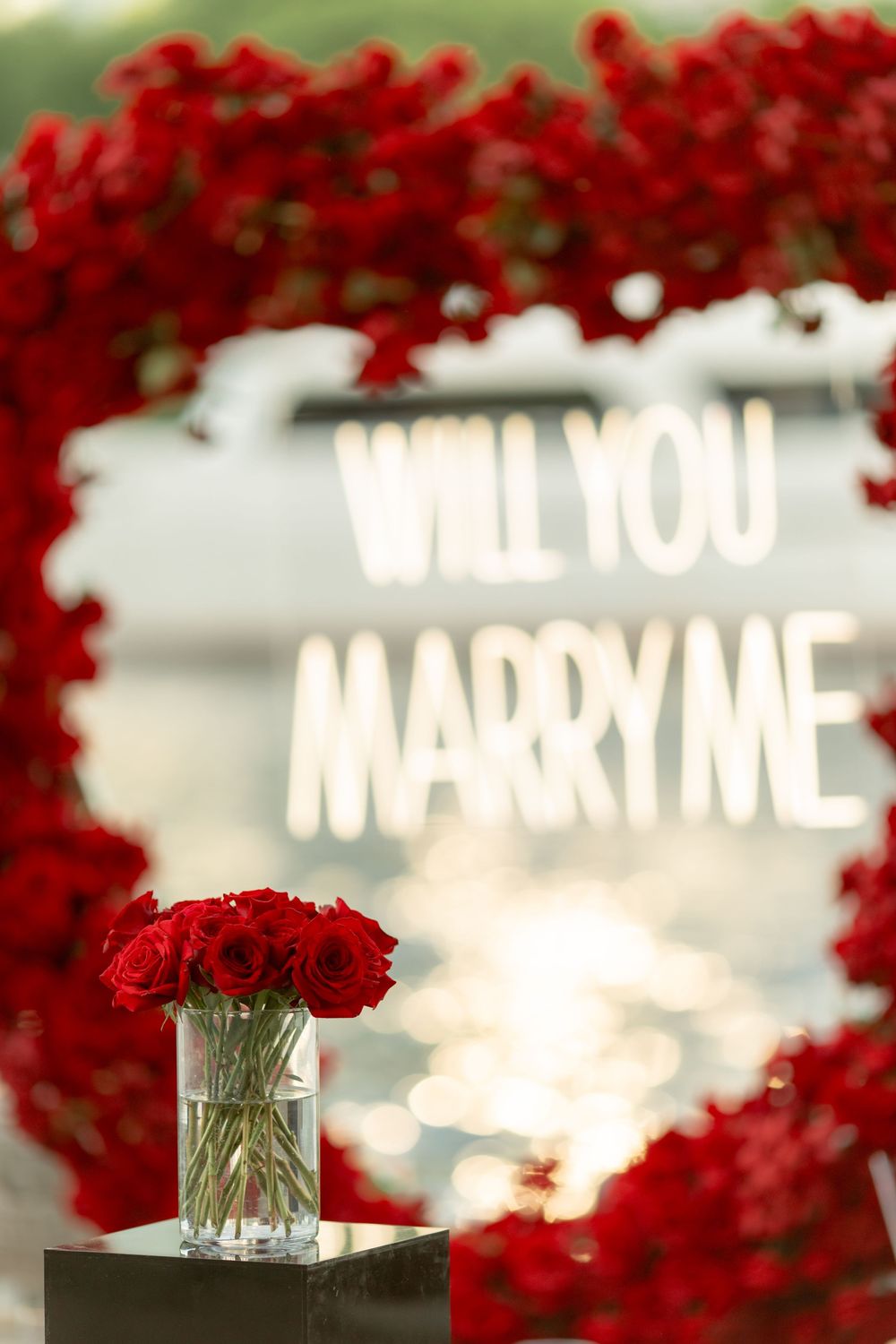 Red rose bouquet in vase framed by heart-shaped floral arch with glowing Will You Marry Me sign.