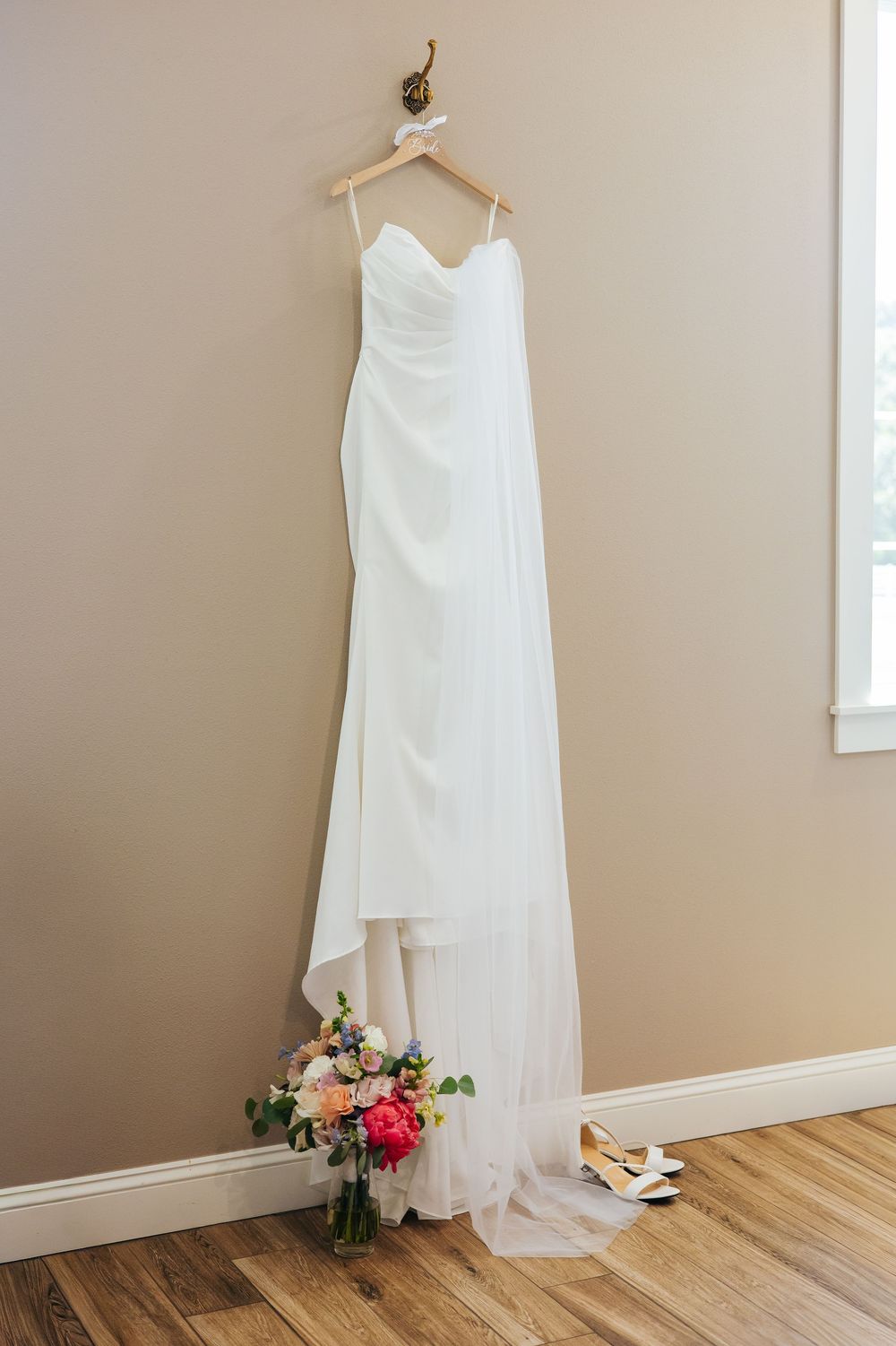 Elegant strapless white wedding gown hanging on wooden hanger with bridal bouquet and heels below.