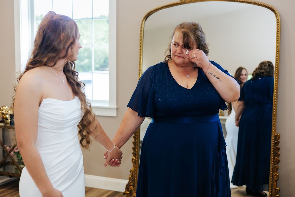 Emotional mother wiping tears while holding hands with bride in white gown before wedding ceremony.
