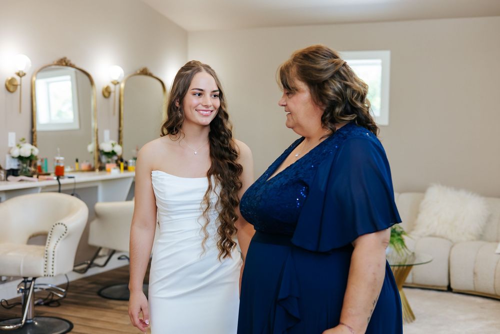 Bride in white strapless gown stands with woman in navy dress in bridal suite.
