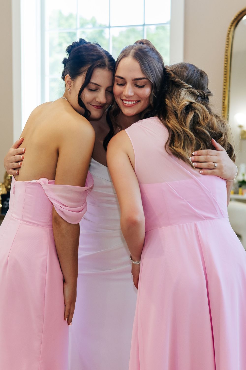 Bride hugging bridesmaids in pink dresses during wedding preparation.
