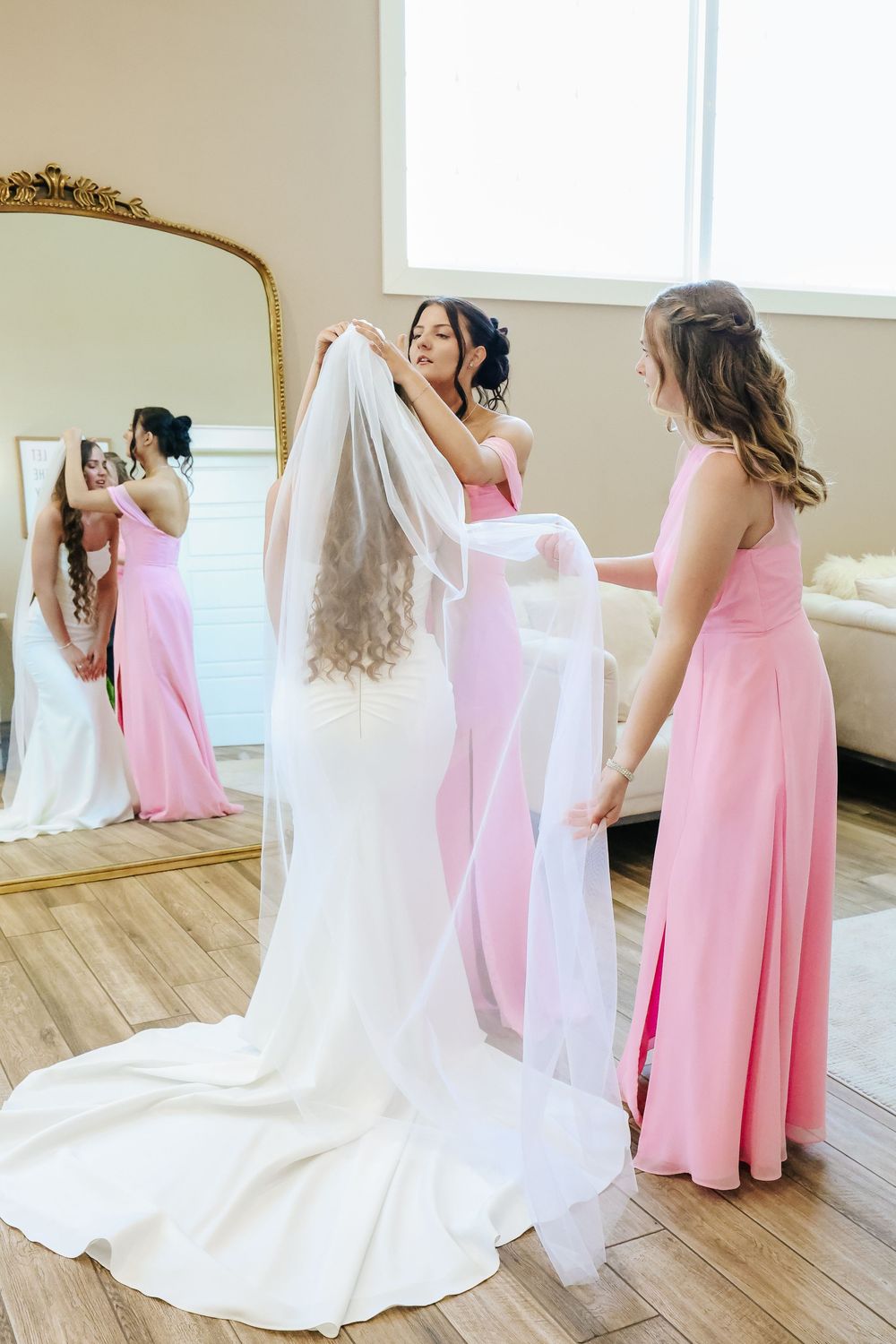Bridesmaids in pink dresses adjusting bride's veil near gold mirror.