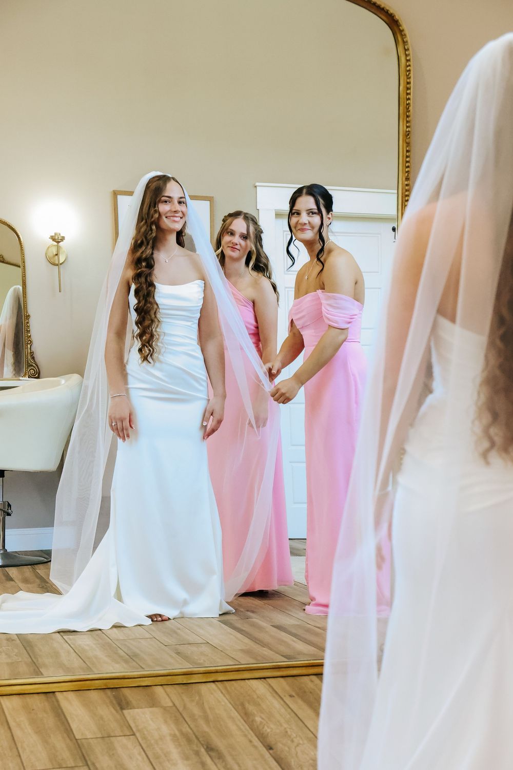 Bride in white gown with veil stands with two bridesmaids in pink dresses reflected in a large mirror.
