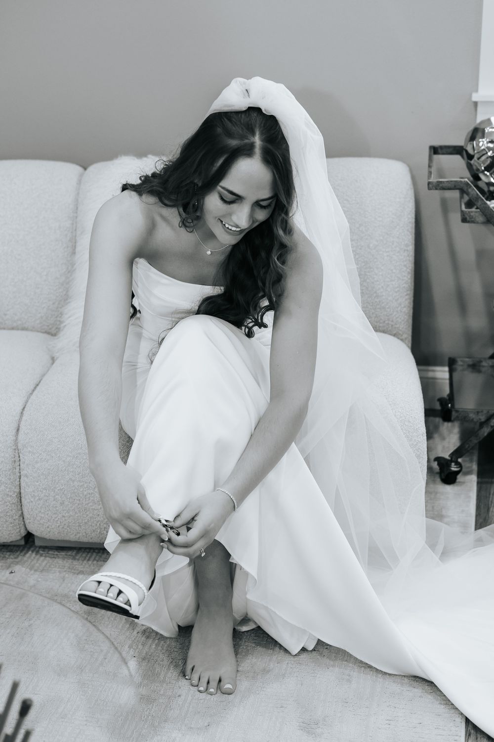 Black and white photo of smiling bride in gown and veil putting on shoes.