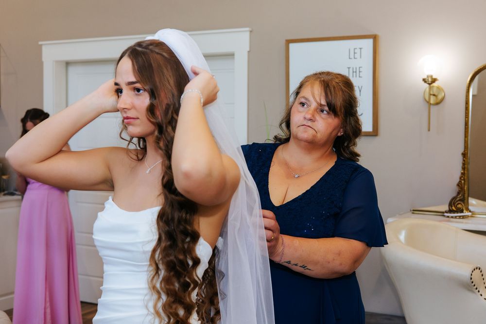 Mother helping bride adjust wedding veil in bridal suite before ceremony.
