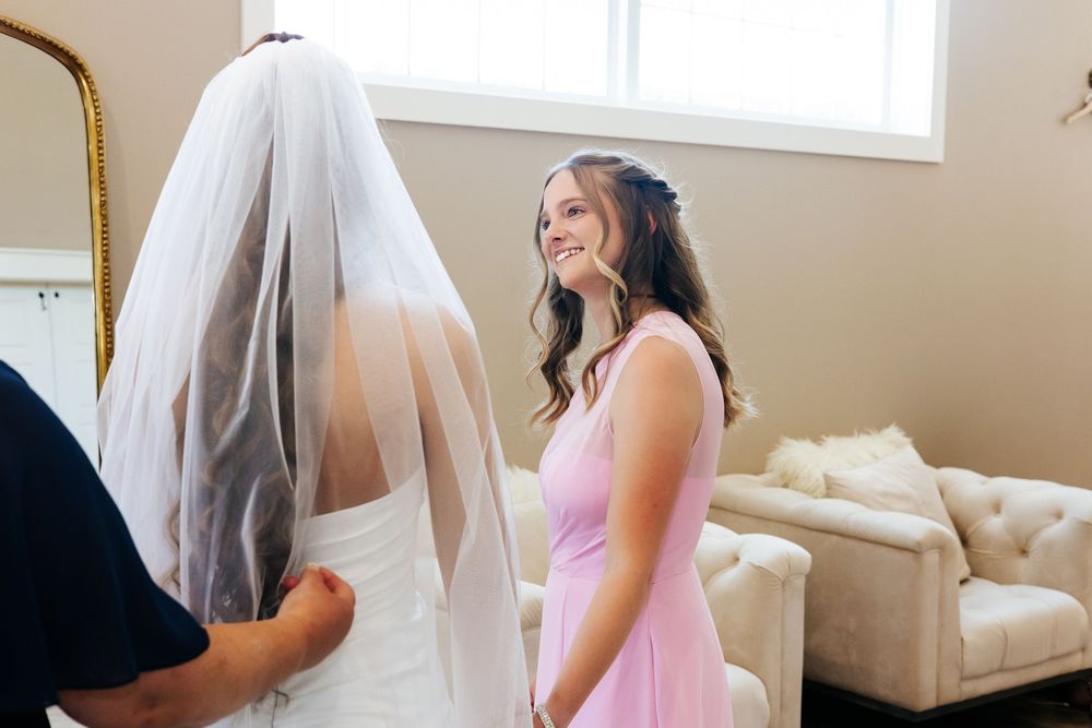 Bridesmaid in pink dress smiling at bride in white gown and veil during wedding preparations.