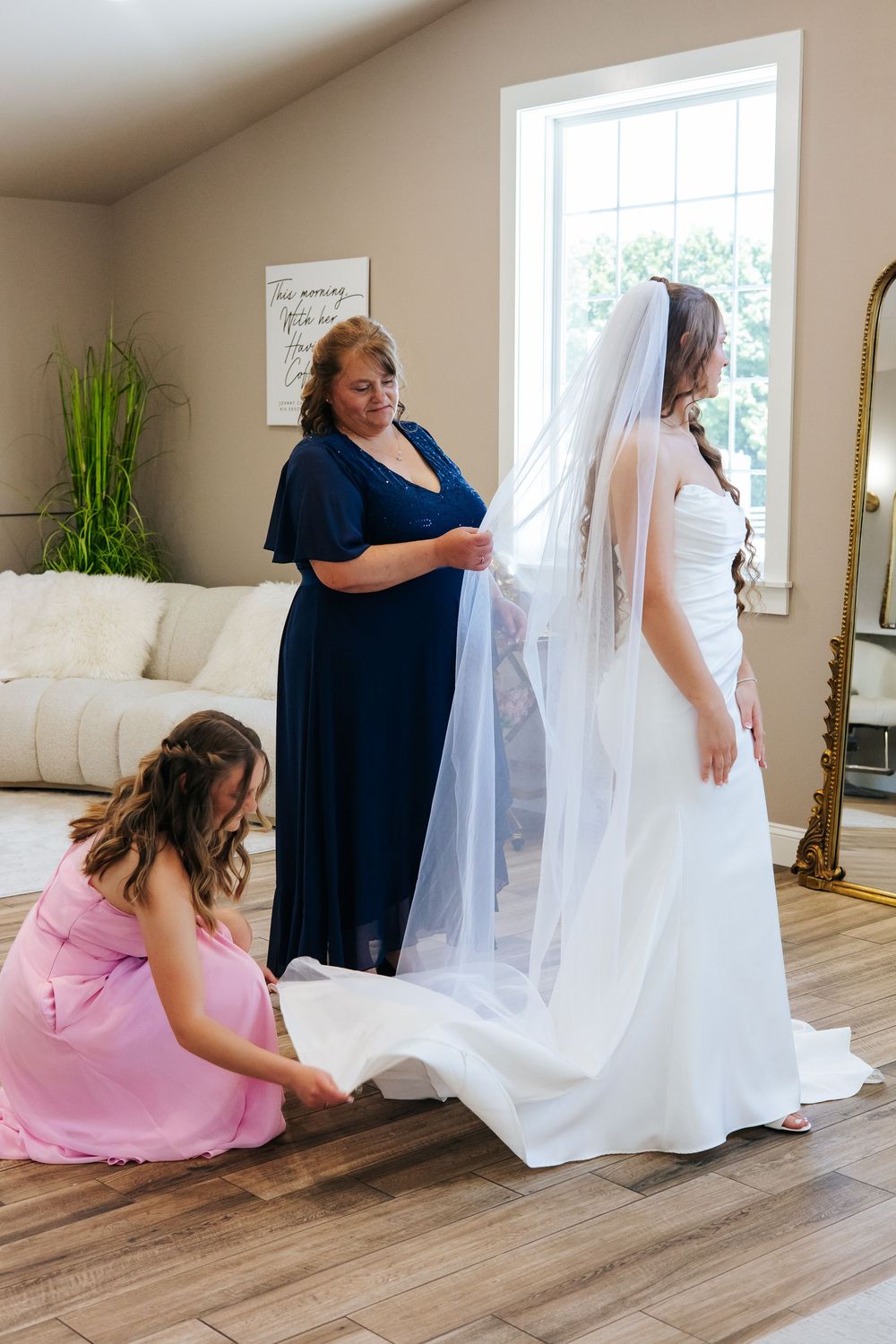 Two women helping bride with white gown and veil in bright dressing room.