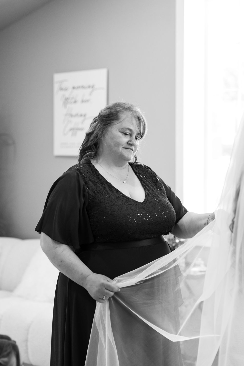 Black and white photo of woman in dark dress holding bridal veil near window.