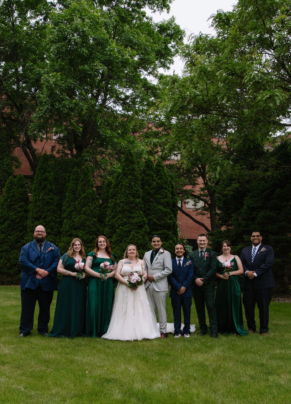 Wedding party spreads out in a line formation on green grass with trees creating a natural backdrop at outdoor celebration.
