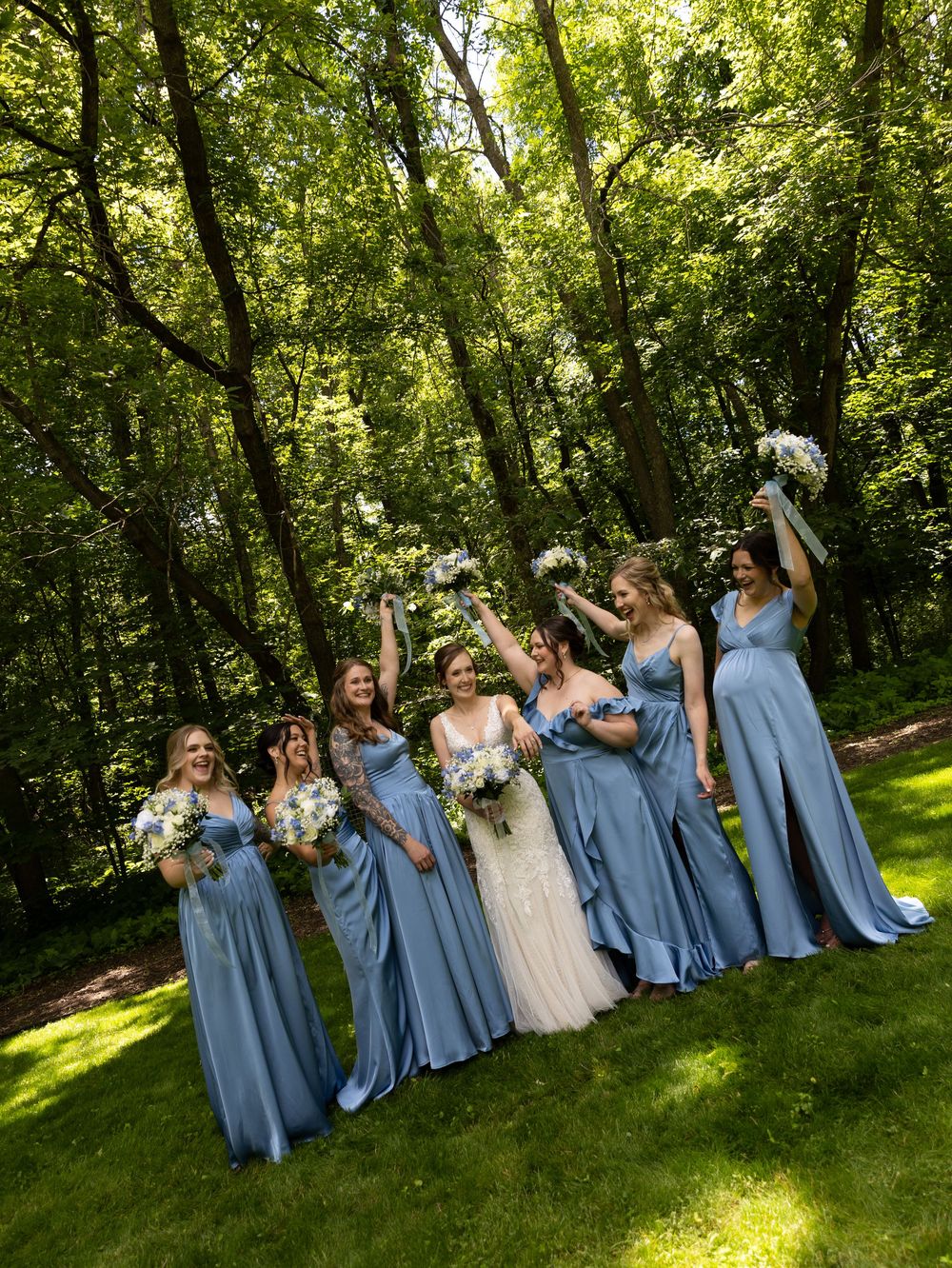 Bridesmaids in blue satin gowns celebrate with bride in white dress, holding bouquets outdoors in lush green forest.