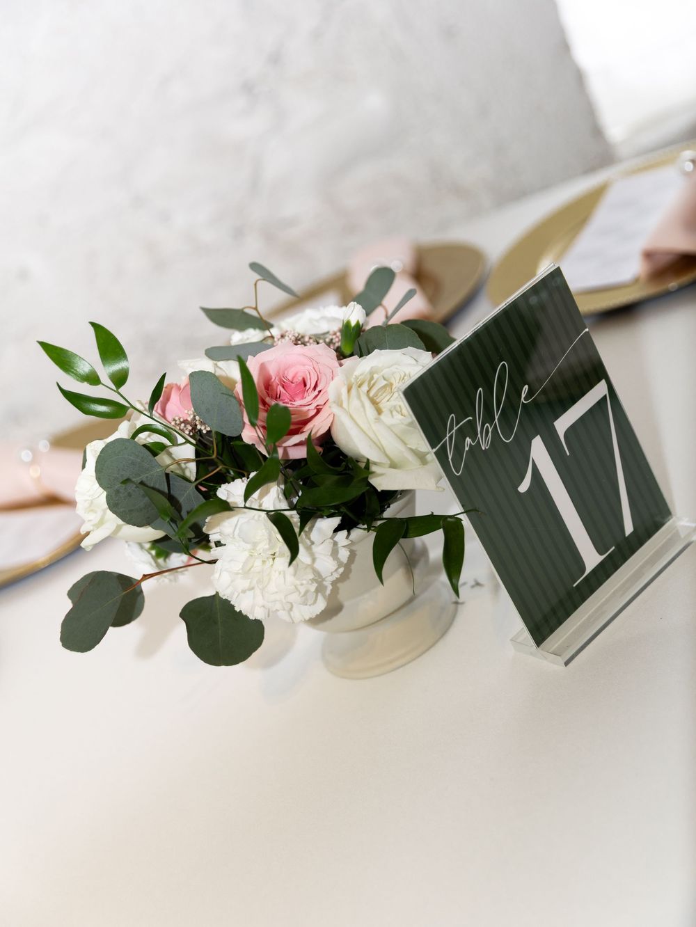 White vase with pink and white roses and eucalyptus next to a dark green table number 17 sign on a white table.
