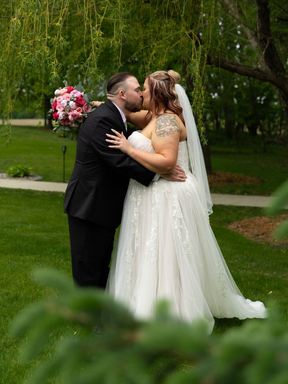 Bride and groom share a romantic kiss outdoors under a willow tree, bride holding pink bouquet in white gown.