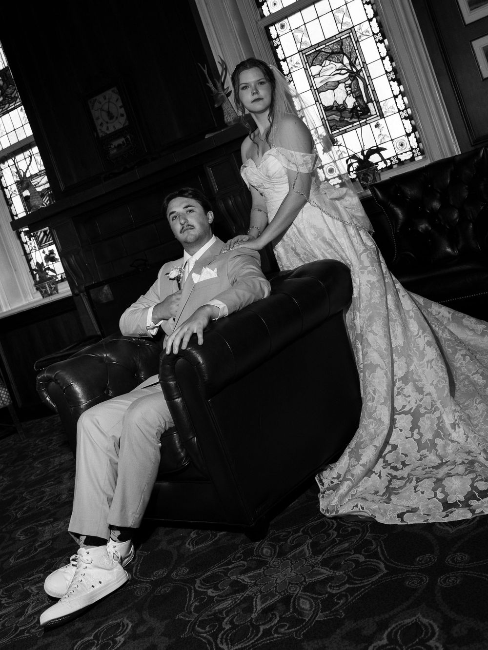 Black and white wedding photo of bride in lace gown and groom in suit seated in leather chair in elegant room.