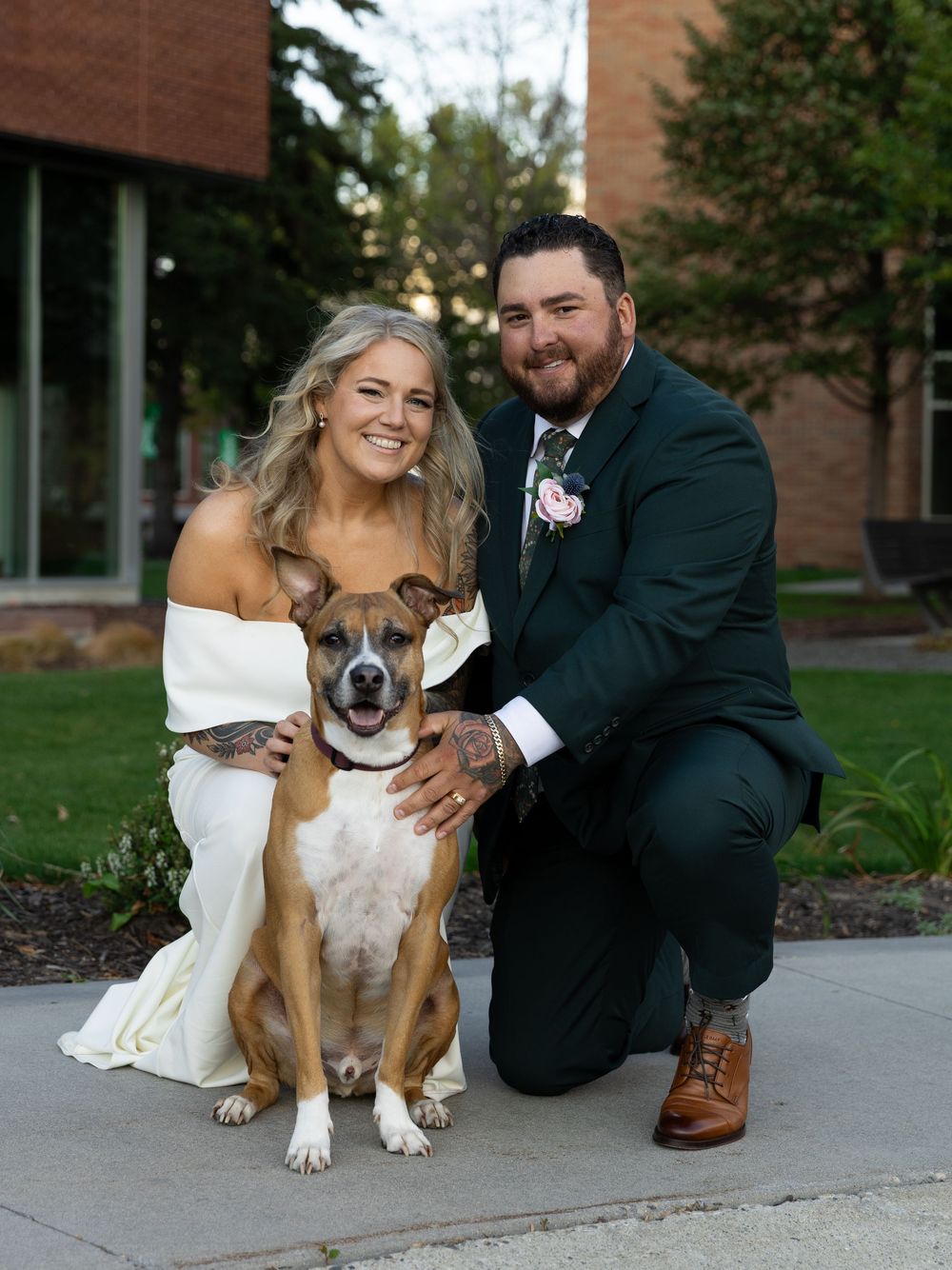 Bride and groom in wedding attire kneel beside their brown and white dog outdoors near a brick building.