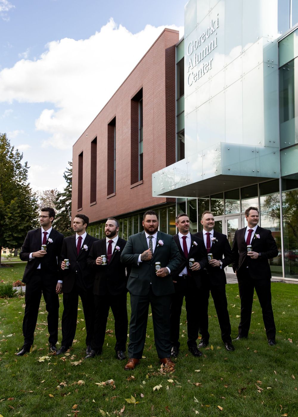 Seven groomsmen in dark suits with burgundy ties pose outside the Cascade Cultural Center on a sunny day.
