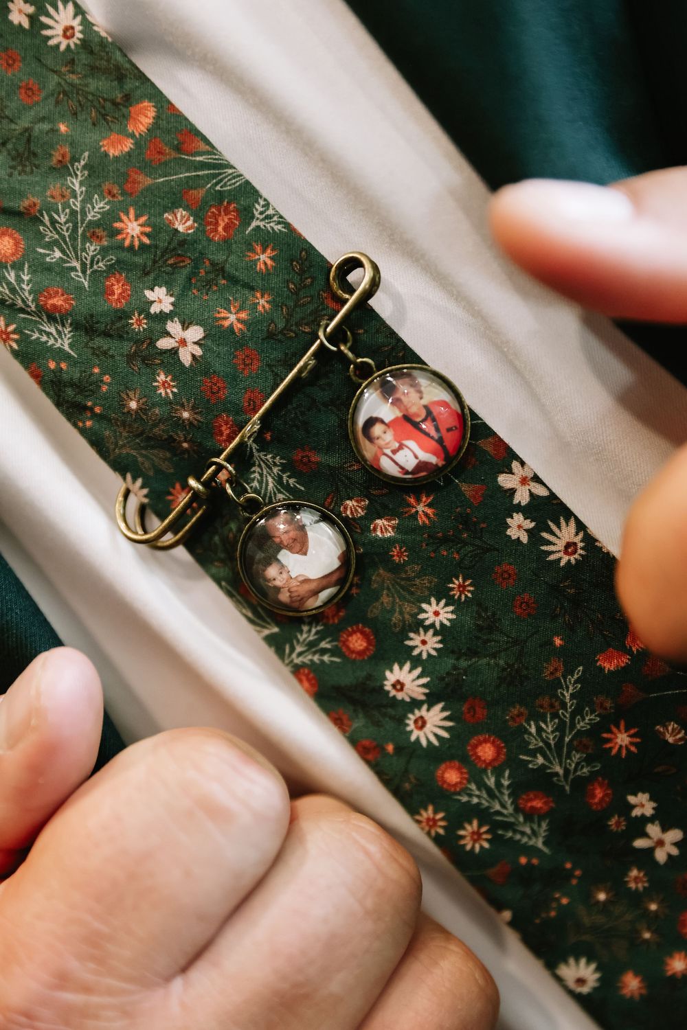 Close-up of a decorative brooch pin with two photo charms on green floral fabric, held by hands.