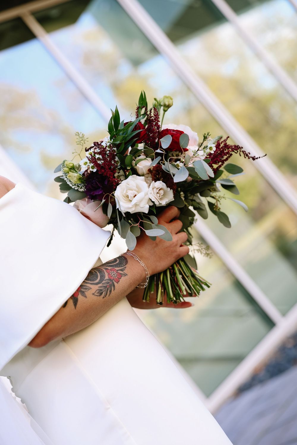 Bride holding a wedding bouquet with white roses, burgundy blooms, and greenery, showcasing a tattooed arm.