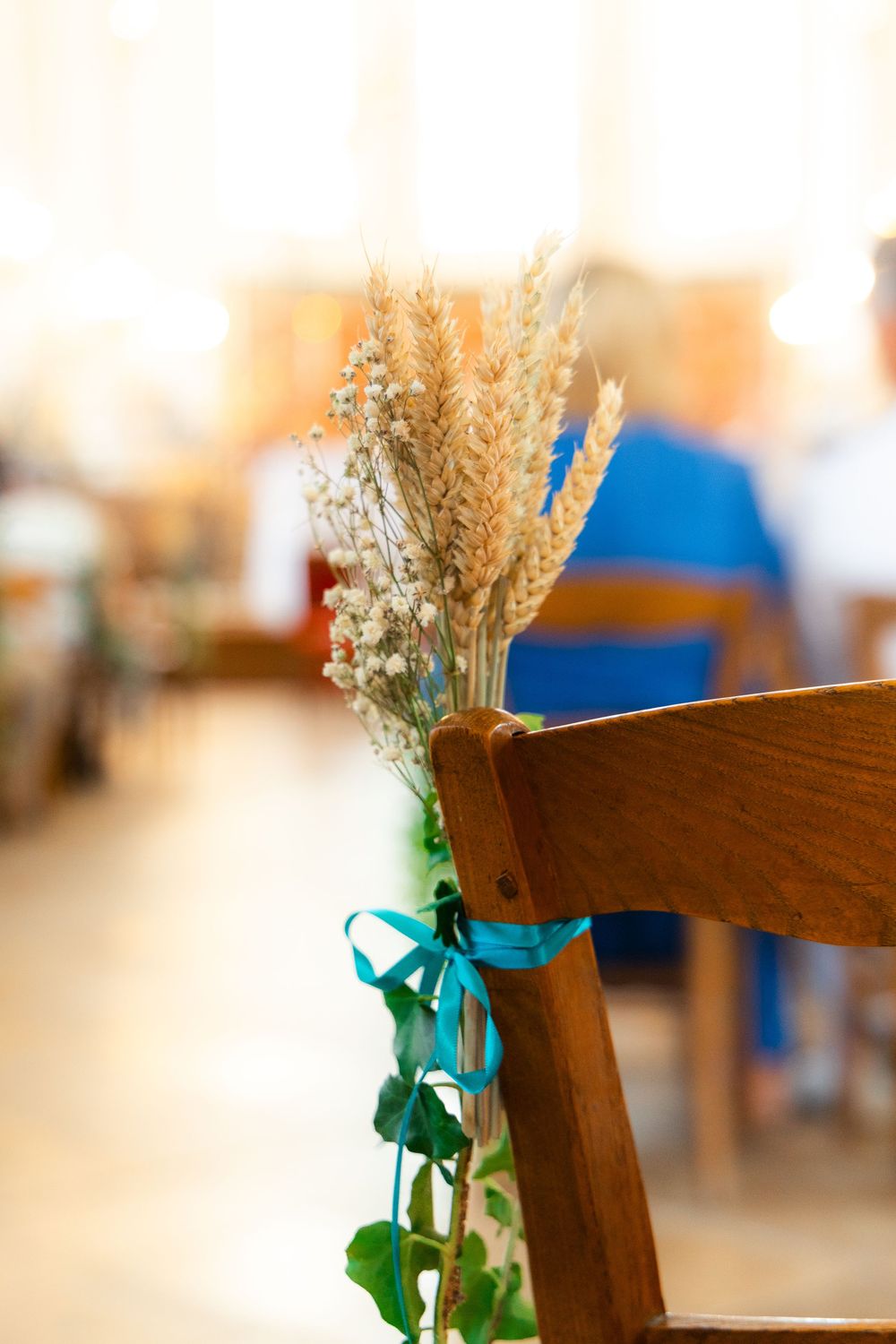 Wooden church chair decorated with dried wheat stalks and teal ribbon bow, elegant wedding ceremony aisle decor.