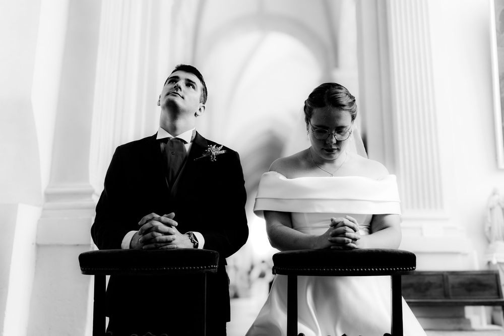 Black and white photo of bride and groom kneeling in prayer during elegant church wedding ceremony.