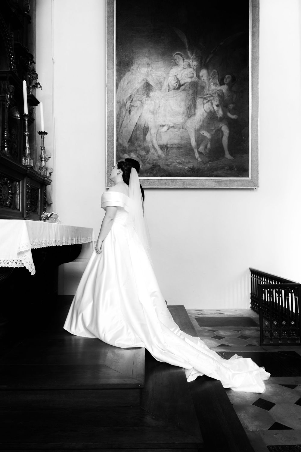 Bride in white gown and veil kneeling in prayer before altar painting in black and white church photo.