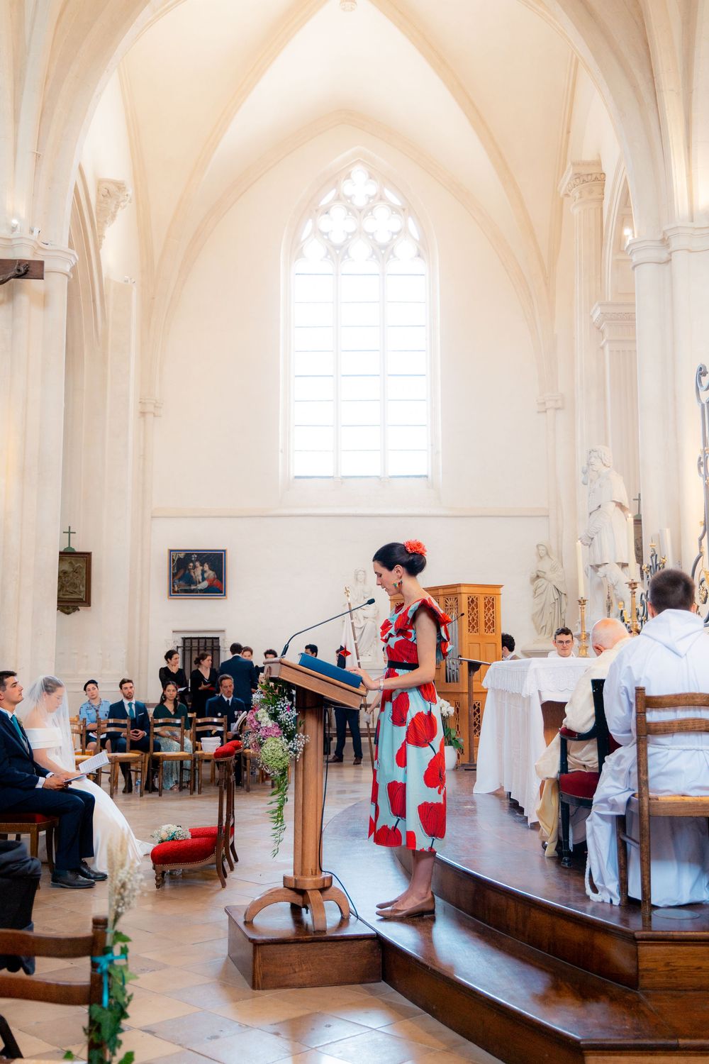 Woman in red floral dress reads at church wedding ceremony podium with gothic arched windows and guests seated in pews.