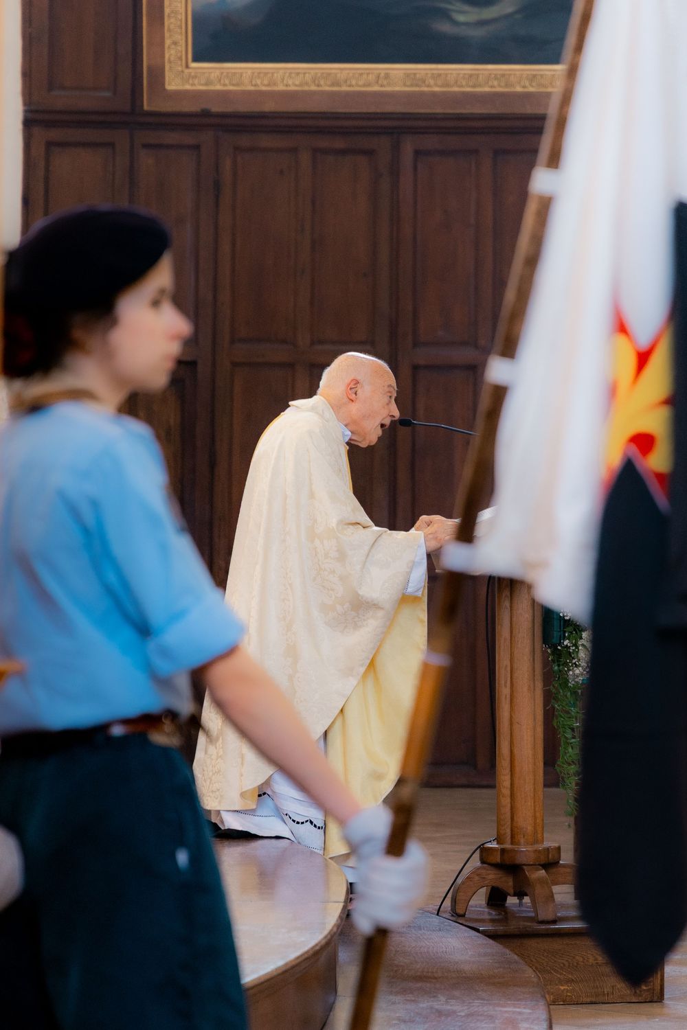 Elderly priest in white vestments performing a religious ceremony inside a church with a young woman holding a flag nearby.