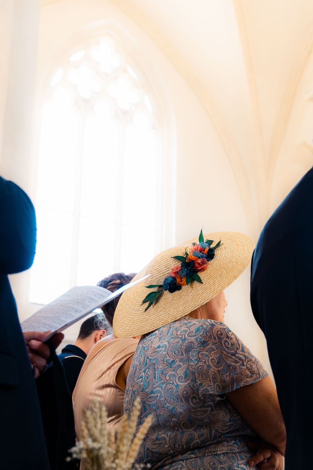 Wedding guest wearing elegant wide-brim straw hat decorated with colorful flowers inside a bright church ceremony.