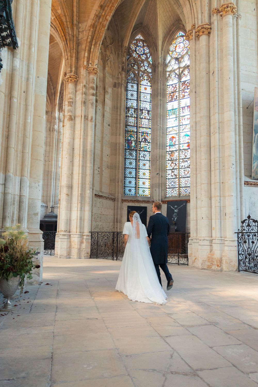 Bride and groom walking hand in hand inside a grand cathedral with stained glass windows and Gothic stone columns.