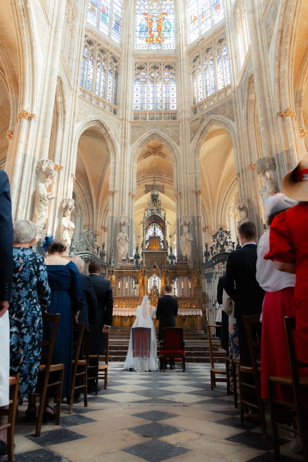 Wedding ceremony in a stunning Gothic cathedral with stained glass windows, bride and groom at the altar surrounded by guests.