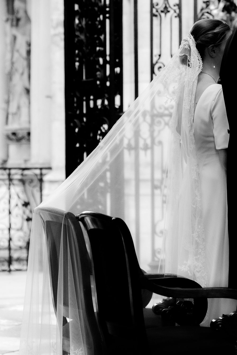 Black and white photo of a bride in a white gown and veil kneeling at a church pew during a wedding ceremony.