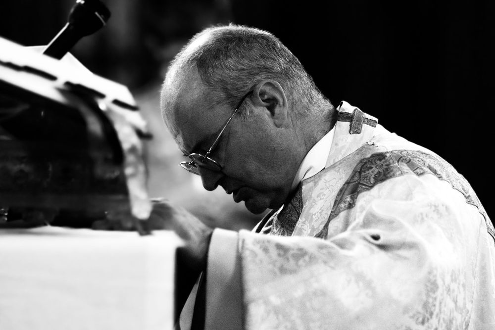Elderly priest in ornate vestments bows head in prayer at altar in black and white photo.