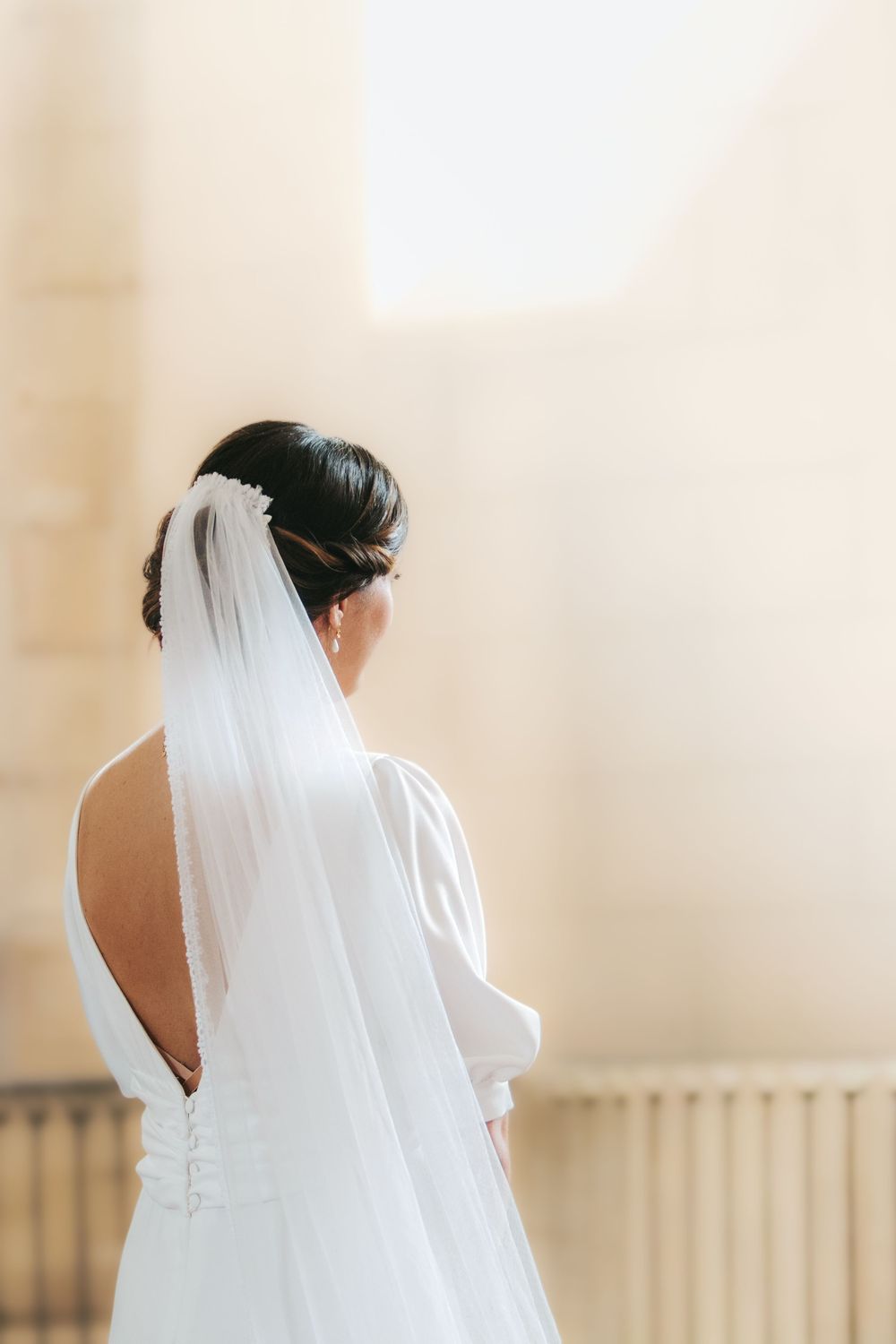 Bride in white backless gown and veil with floral hairpiece, viewed from behind in soft natural light.