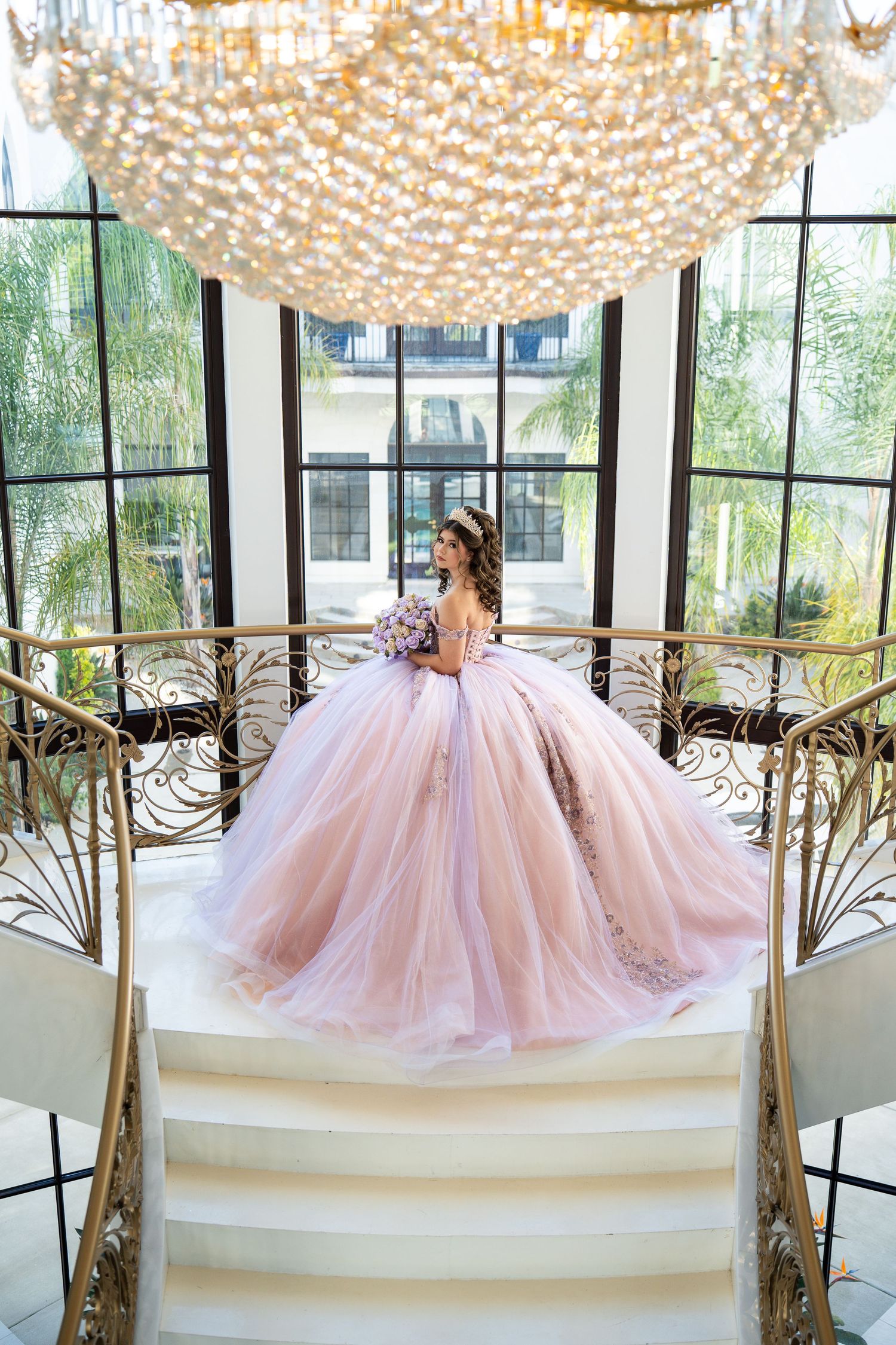 Young woman in a pink ball gown quinceañera dress stands on grand staircase beneath crystal chandelier.