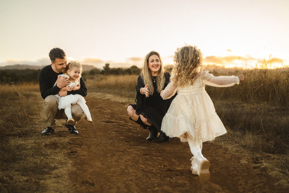 Family enjoying golden hour sunset on a dirt path, parents playing with two young daughters in a field.