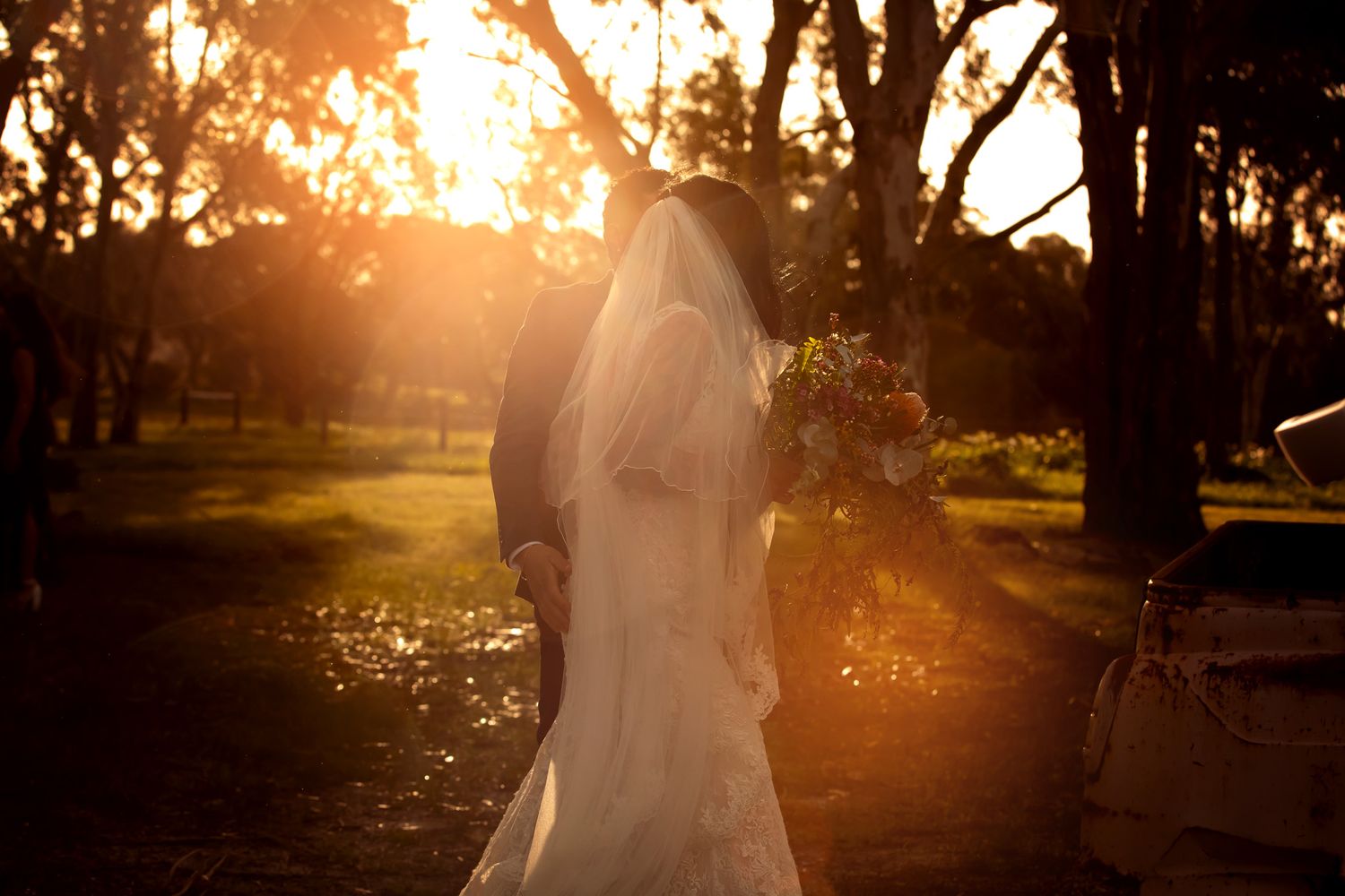 Bride and groom silhouetted against golden sunset light in a tree-lined park on their wedding day.