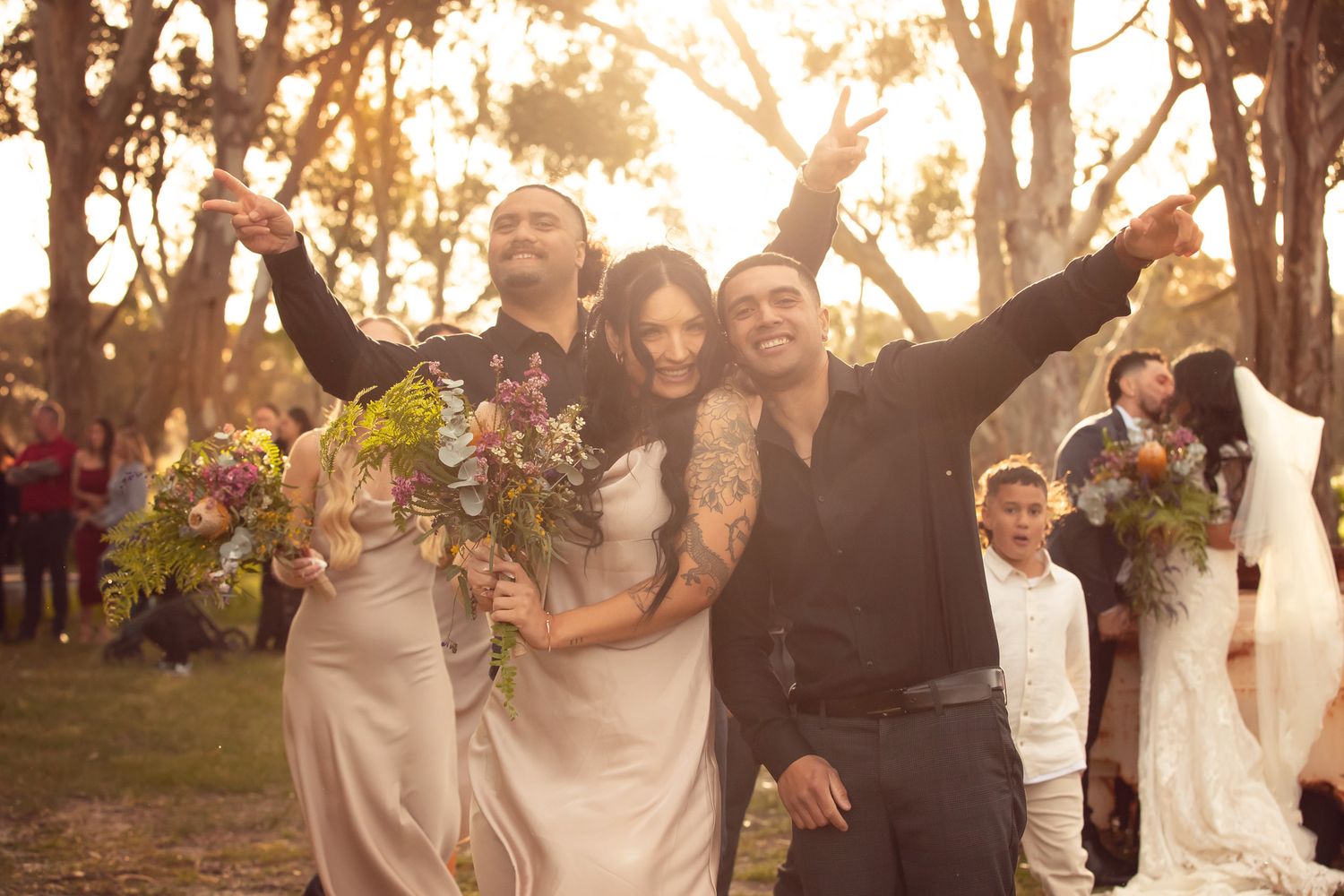 Joyful wedding party celebrating outdoors with bouquets raised, golden sunlight filtering through trees at a garden ceremony.