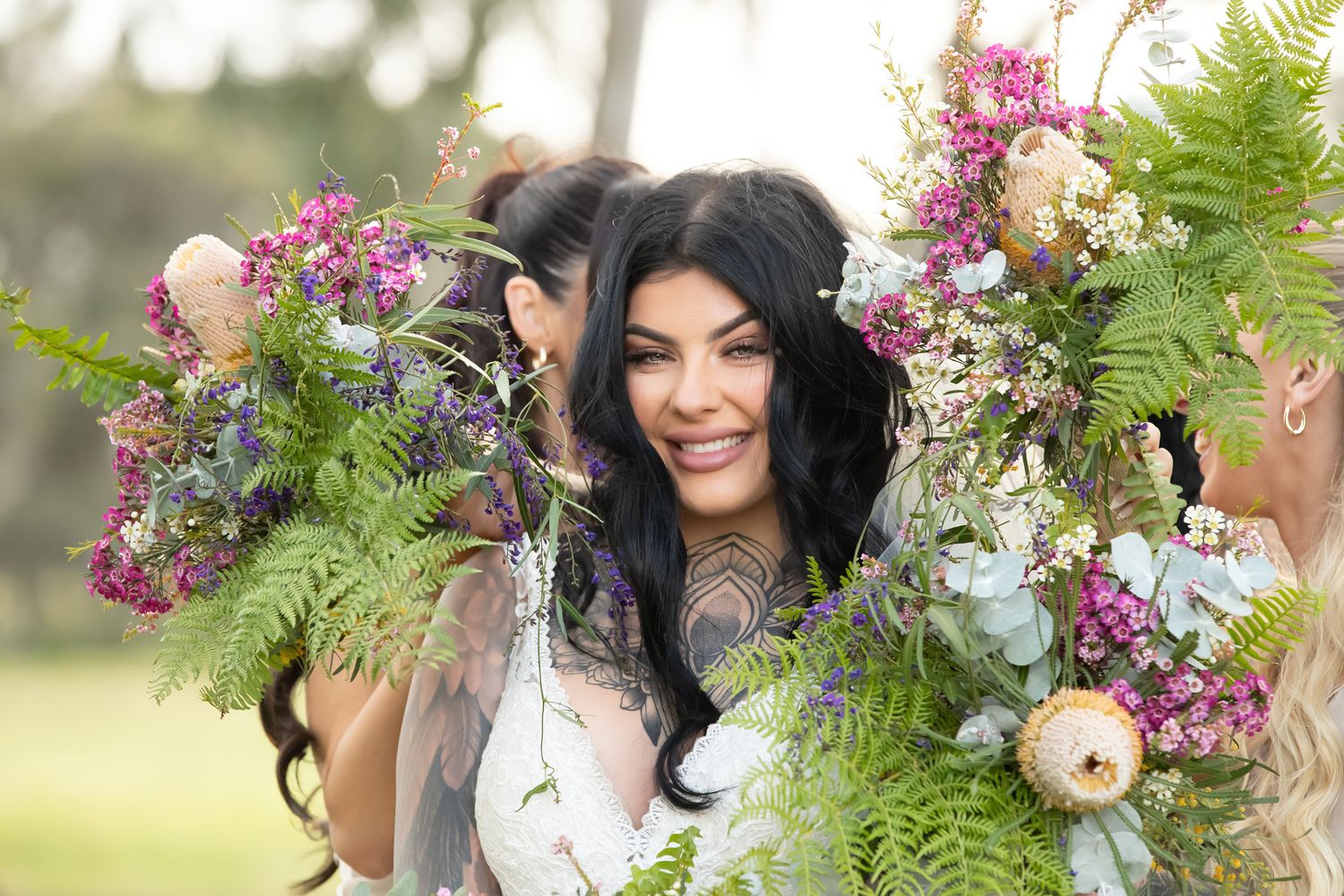 Smiling woman with long dark hair surrounded by a lush floral wreath of wildflowers and ferns.