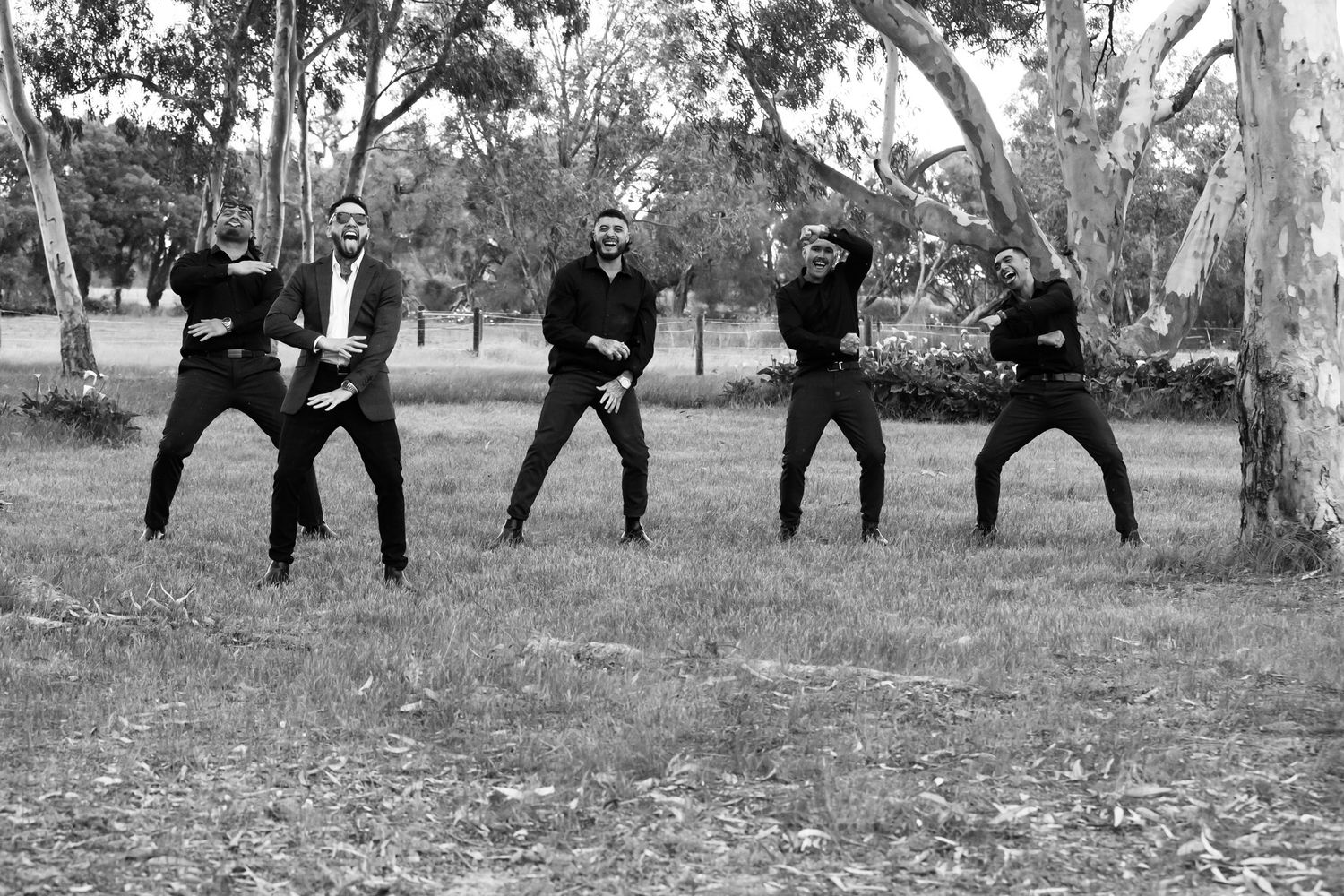 Five men in black suits dancing outdoors in a park among eucalyptus trees in a fun black and white photo.