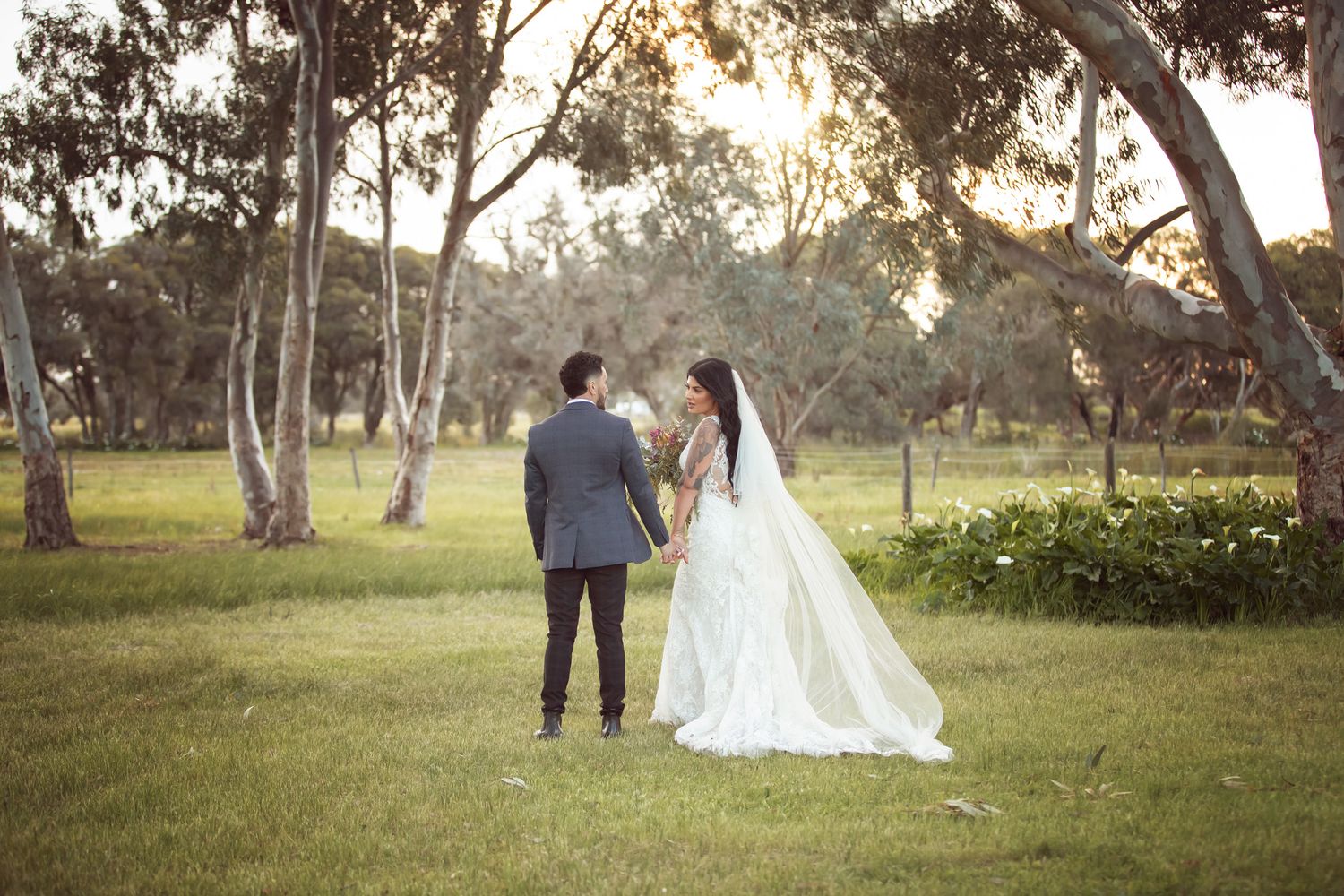 A bride and groom stand together in a lush green park surrounded by tall trees during their outdoor wedding ceremony.