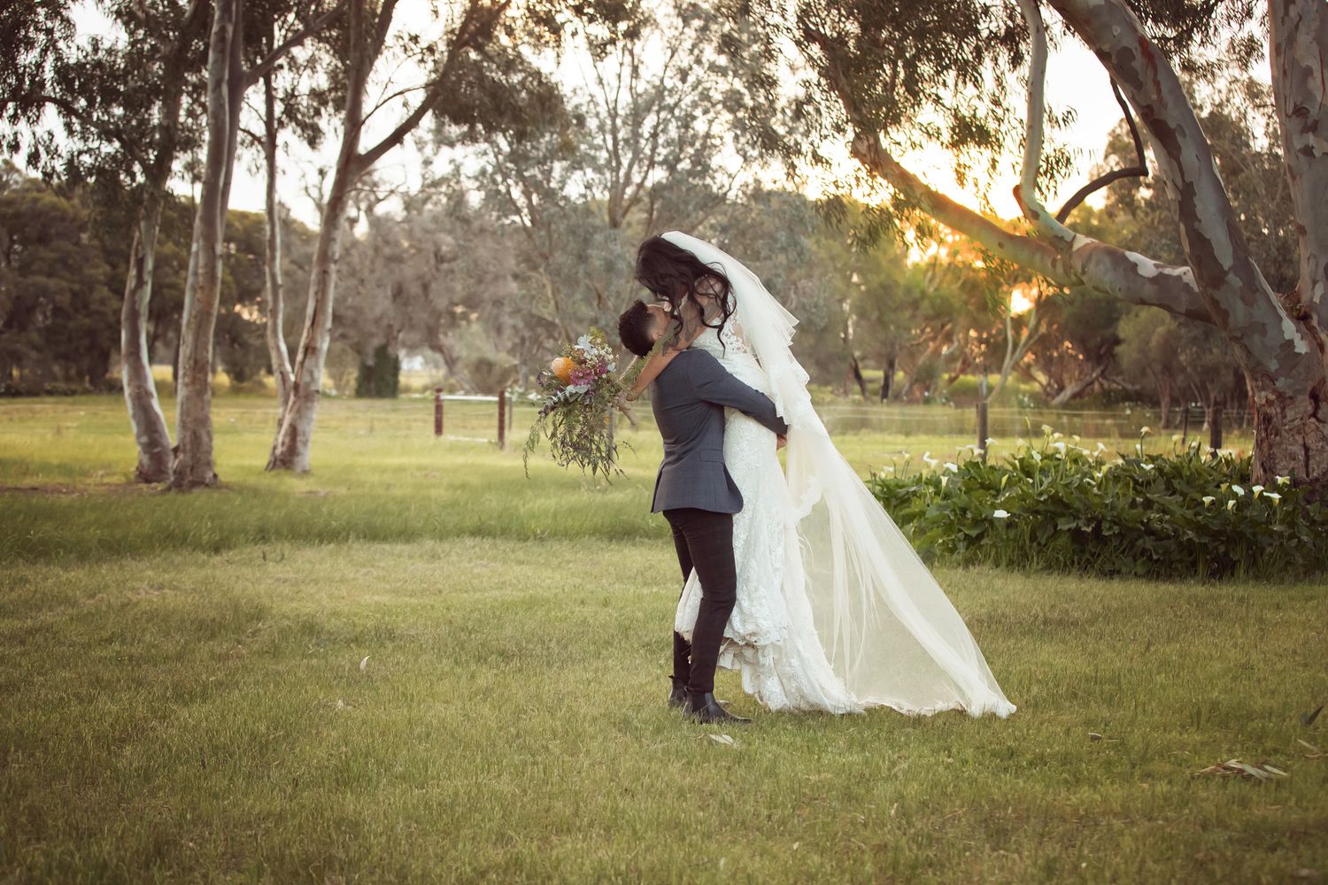 Bride and groom share a romantic kiss at sunset in a lush green park, with the bride in a flowing white gown.