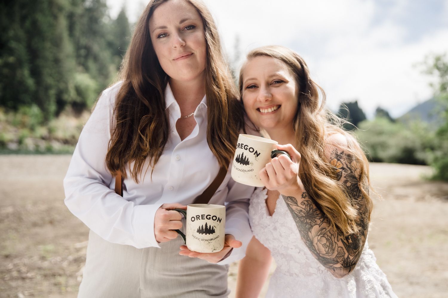 Two brides in white holding Oregon mugs smile together outdoors at their wedding celebration.