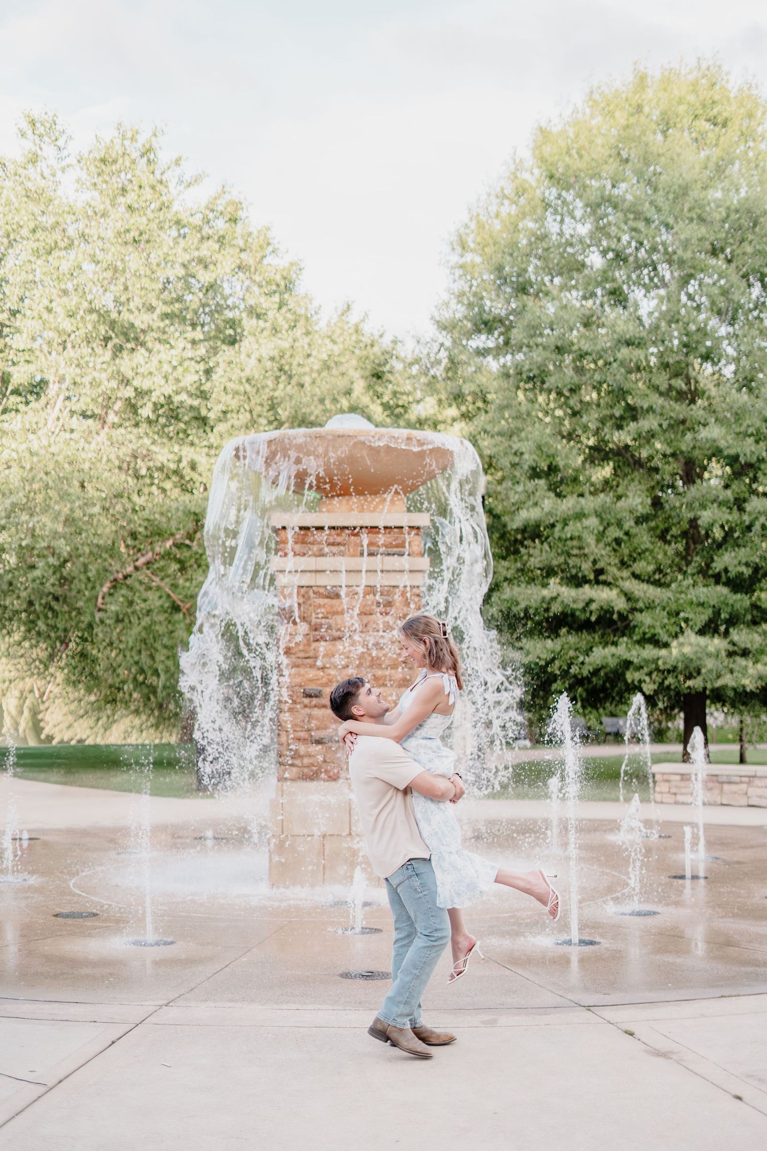 Couple embracing by a fountain in a park, man lifting woman as water cascades behind them on a sunny day.