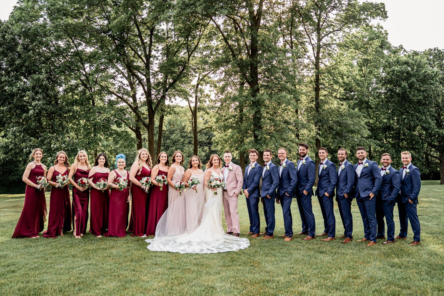 Large wedding party posing outdoors, bridesmaids in burgundy gowns, groomsmen in navy suits, bride and groom at center.