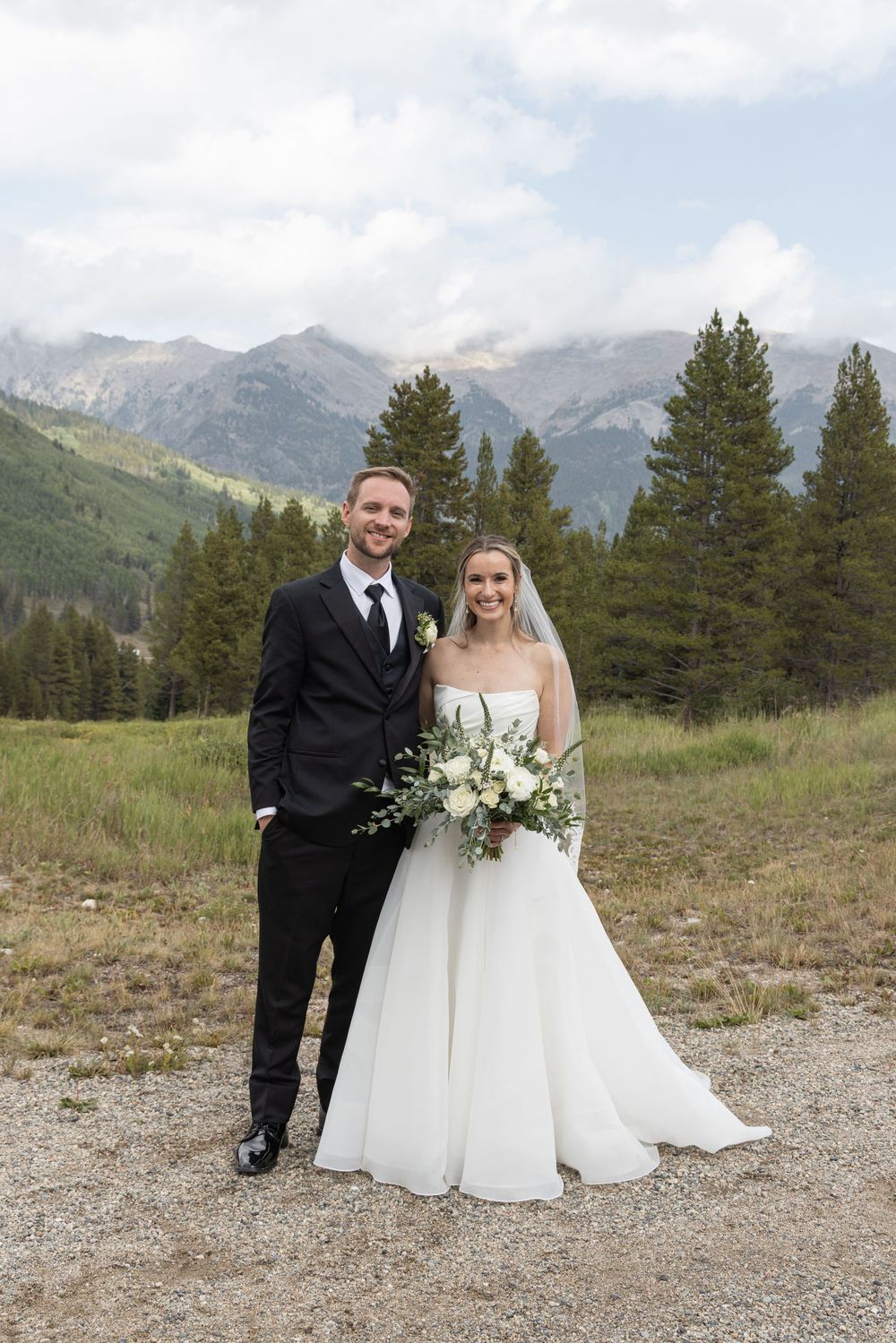 Bride and groom posing together in mountain meadow, bride holding white bouquet, evergreen trees and rocky peaks behind them.