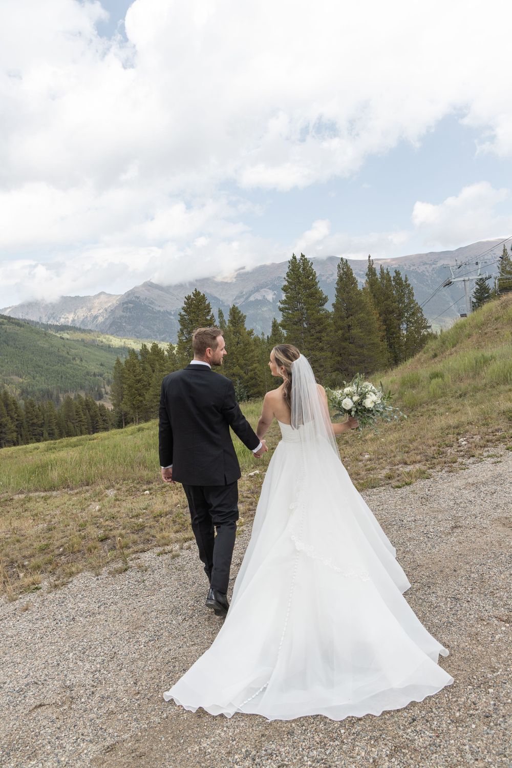 A bride and groom walk hand-in-hand along a mountain path surrounded by pine trees and scenic peaks.